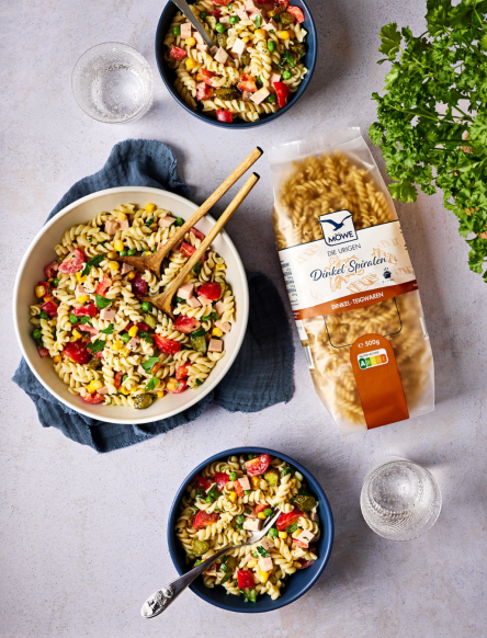 Schüssel Nudeln mit zwei Schalen, eine Packung Dinkel-Spiralen und verschiedene Zutaten für einen Salat. Siehe Rezept.