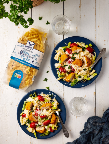 Eine Packung Möwe Meine Liebsten Spiralen auf weißem Holzuntergrund, serviert auf zwei Tellern dann Nudeln mit Salat und Chicken Nuggets als Caesar Salad Alternative.