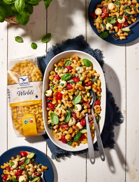 Eine Packung Möwe Wirbli Nudeln links im Bild. Daneben eine Auflaufform, die einen knusprigen Nudelsalat zeigt. Darüber und darunter jeweils ein Teller. Siehe Rezept.