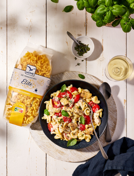 Eine Packung Möwe Die Himmlischen Elite Nudeln auf hellem Holzuntergrund. Daneben ein Teller mit Nudeln, Tomaten und Fisch. Siehe Rezept.