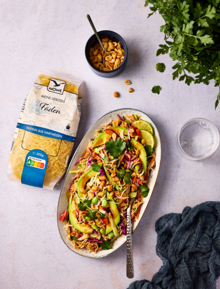 Eine Packung Möwe Meine Liebsten Faden-Nudeln auf hellgrauem Untergrund. Daneben ein oval geformter Teller, auf dem Erdnüsse, Avocado, Karotten, Limettenscheiben mit den Nudeln gemischt sind. Siehe Rezept.