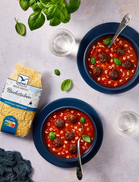 Eine Packung Möwe Buchstaben Nudeln links im Bild. Daneben zwei Schüsseln mit Tomatensuppe und Fleischbällchen, in denen die Buchstaben schwimmen.