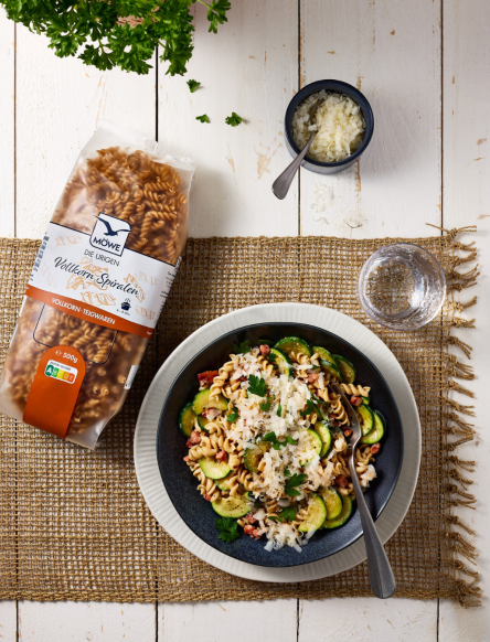 Eine Packung Möwe Vollkorn Spiralen. Daneben ein Teller mit einem Gericht aus den Spiralen und Zucchini und Speck mit Parmesan garniert. Siehe Rezept.