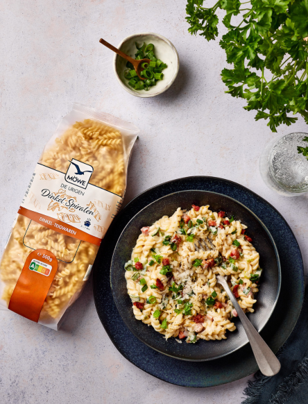 Die Urigen Dinkel Spiralen der Marke Möwe liegen links im Bild, daneben ein dunkler Teller, auf dem gekochte Nudeln mit Flammkuchen Soße drapiert sind.