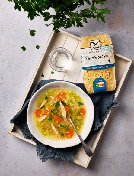 Ein hellgrauer Untergrund, darauf ein weißes Tablett in Holzoptik. Darauf wiederum ein Gericht angerichtet: Ein Teller Hühnersuppe. Darüber eine Packung Buchstabennudeln der Marke Möwe.