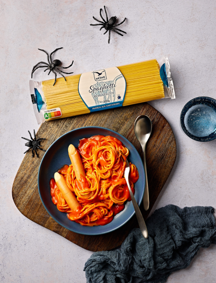 Ein hellgrauer Untergrund, auf dem ein Halloween-Essen serviert ist: Ein Teller mit Spaghetti in roter Tomatensoße, dazu Würstchen als Finger angerichtet. Darüber eine Packung Spaghetti der Marke Möwe sowie dekorative Plastik-Spinnen.