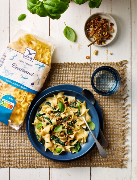Ein weißer Holzuntergrund, auf dem ein aus Jute gewebtes Tischset liegt. Darauf ein Teller Nudeln mit Spinat-Gorgonzola Soße. Daneben eine Packung Breitband Nudeln der Marke Möwe.