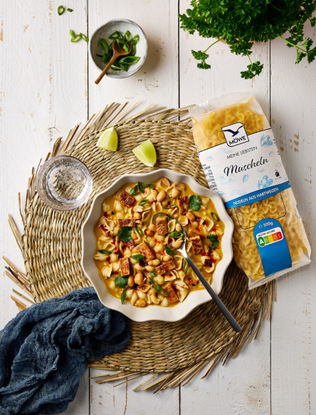 Ein heller weißer Holztisch, auf dem Essen angerichtet ist. Auf einem Jute-Untersetzer ist ein Teller Thai-Nudelsuppe zu sehen mit Muschelnudeln. Daneben liegt eine Packung der Marke Möwe Meine Liebsten Muscheln.