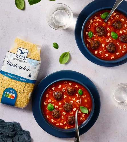 Eine Packung Möwe Buchstaben Nudeln links im Bild. Daneben zwei Schüsseln mit Tomatensuppe und Fleischbällchen, in denen die Buchstaben schwimmen.
