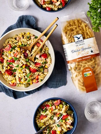 Schüssel Nudeln mit zwei Schalen, eine Packung Dinkel-Spiralen und verschiedene Zutaten für einen Salat. Siehe Rezept.