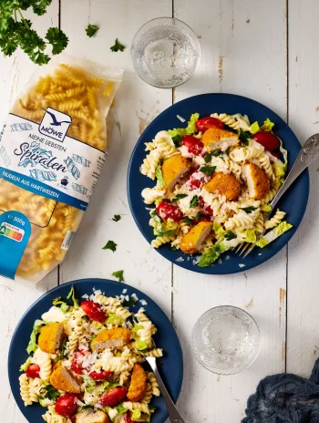 Eine Packung Möwe Meine Liebsten Spiralen auf weißem Holzuntergrund, serviert auf zwei Tellern dann Nudeln mit Salat und Chicken Nuggets als Caesar Salad Alternative.