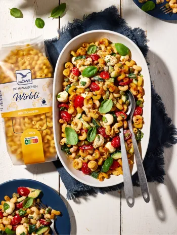 Eine Packung Möwe Wirbli Nudeln links im Bild. Daneben eine Auflaufform, die einen knusprigen Nudelsalat zeigt. Darüber und darunter jeweils ein Teller. Siehe Rezept.
