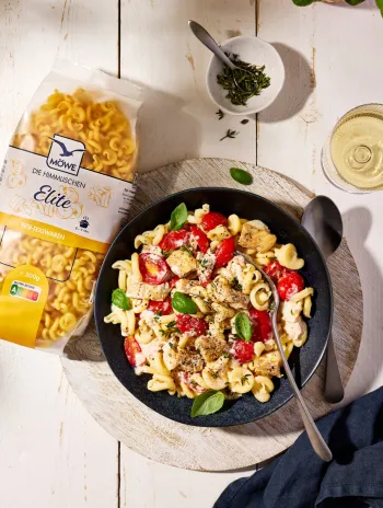 Eine Packung Möwe Die Himmlischen Elite Nudeln auf hellem Holzuntergrund. Daneben ein Teller mit Nudeln, Tomaten und Fisch. Siehe Rezept.