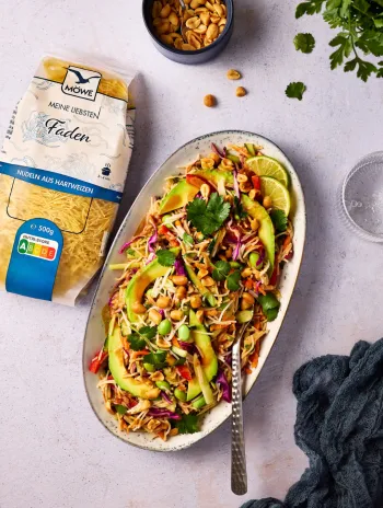 Eine Packung Möwe Meine Liebsten Faden-Nudeln auf hellgrauem Untergrund. Daneben ein oval geformter Teller, auf dem Erdnüsse, Avocado, Karotten, Limettenscheiben mit den Nudeln gemischt sind. Siehe Rezept.