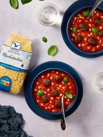 Eine Packung Möwe Buchstaben Nudeln links im Bild. Daneben zwei Schüsseln mit Tomatensuppe und Fleischbällchen, in denen die Buchstaben schwimmen.