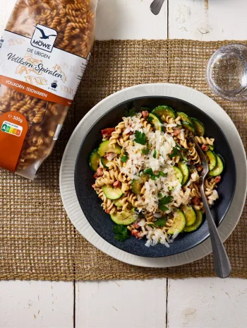 Eine Packung Möwe Vollkorn Spiralen. Daneben ein Teller mit einem Gericht aus den Spiralen und Zucchini und Speck mit Parmesan garniert. Siehe Rezept.