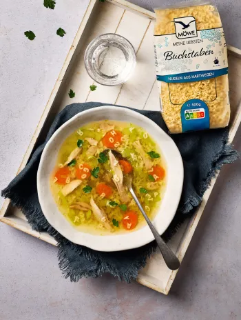 Ein hellgrauer Untergrund, darauf ein weißes Tablett in Holzoptik. Darauf wiederum ein Gericht angerichtet: Ein Teller Hühnersuppe. Darüber eine Packung Buchstabennudeln der Marke Möwe.