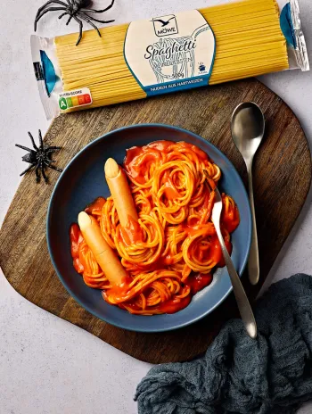 Ein hellgrauer Untergrund, auf dem ein Halloween-Essen serviert ist: Ein Teller mit Spaghetti in roter Tomatensoße, dazu Würstchen als Finger angerichtet. Darüber eine Packung Spaghetti der Marke Möwe sowie dekorative Plastik-Spinnen.
