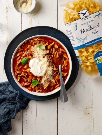 Ein heller Holztisch, auf dem ein Essen serviert ist: Ein Teller mit Tomatensuppe, in der Nudeln schwimmen und der mit einem Klecks Schmand verziert ist. Daneben eine Packung Nudeln der Marke Möwe Meine Liebsten Breitband. Darüber noch ein Schälchen Parmesan.