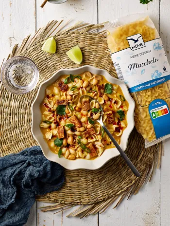 Ein heller weißer Holztisch, auf dem Essen angerichtet ist. Auf einem Jute-Untersetzer ist ein Teller Thai-Nudelsuppe zu sehen mit Muschelnudeln. Daneben liegt eine Packung der Marke Möwe Meine Liebsten Muscheln.
