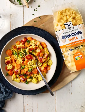 Ein heller Holztisch, auf dem ein Gericht platziert ist. Man erkennt Nudeln in Basketball-Form und Karotten-Scheiben. Daneben eine Packung der SEAWOLVES-PASTA der Marke Möwe.